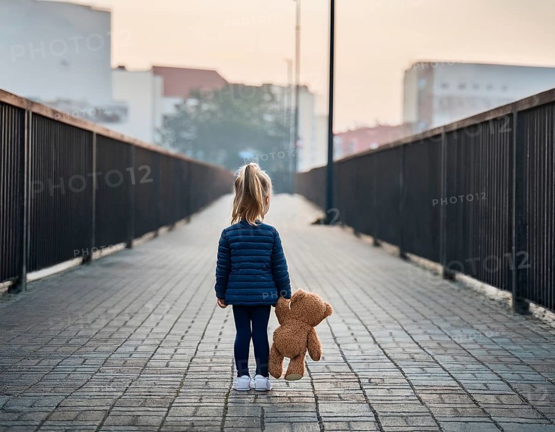 A young child stands alone holding a teddy bear in an urban outdoor setting, symbolizing innocence and isolation, AI generated