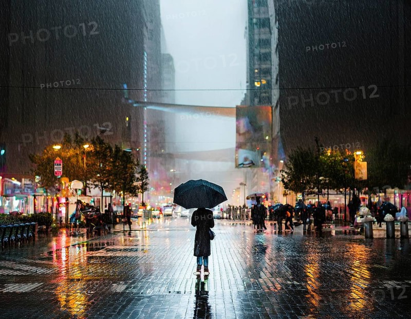 A solitary figure with an umbrella stands amidst a bustling city illuminated by lights, with reflections on the rain-soaked pavement creating a vibrant and atmospheric urban evening scene, AI generated