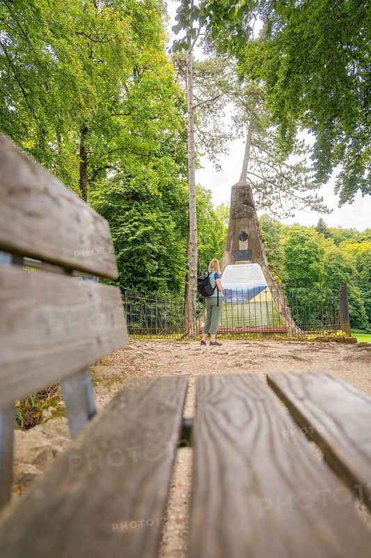 Woman with rucksack looking at a monument in the forest, near a wooden bench, Lichtenstein Castle, Lichtenstein, Swabian Alb, Germany
