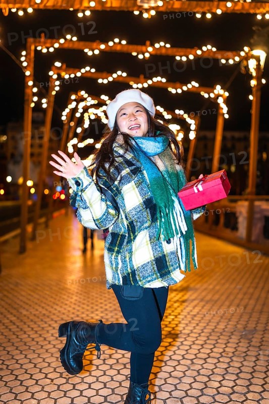 Happy young woman holding a christmas gift, reveling in the enchanting glow of festive lights and decorations on a winter night