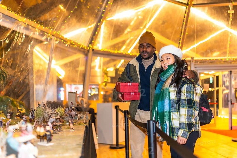 Multiethnic couple holding christmas gifts looking at a nativity scene in a christmas village illuminated at night