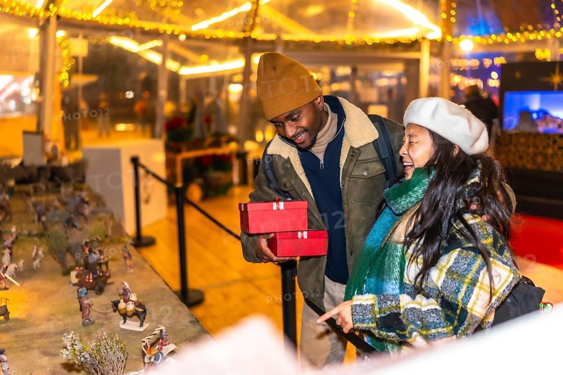 Young multiethnic couple holding christmas gifts admiring a nativity scene at a christmas market, enjoying winter holidays together