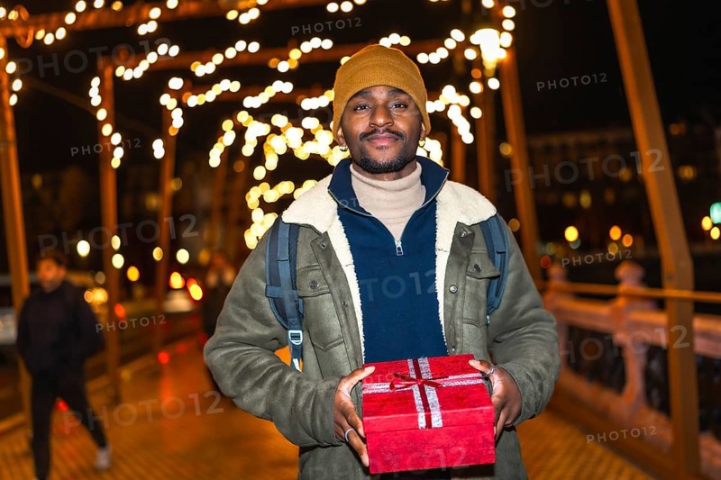 Young man holding christmas gift while walking on a city bridge decorated with christmas lights