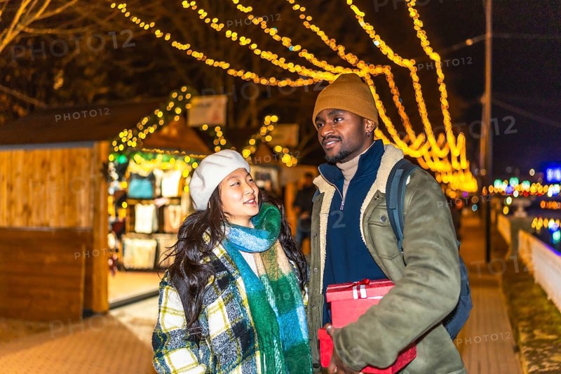 Young multiethnic couple walking and enjoying christmastime at a decorated outdoor market with festive lights