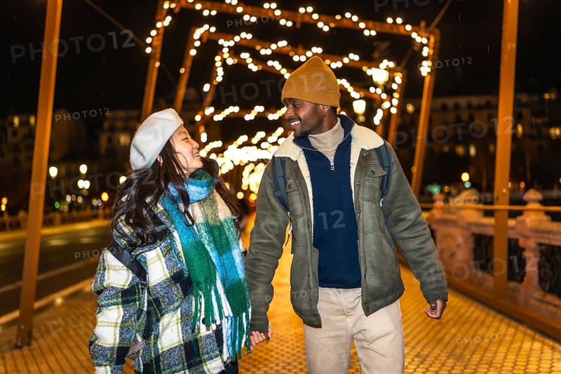 Young multiethnic tourists couple holding hands, enjoying christmas lights decoration at night while walking on a bridge
