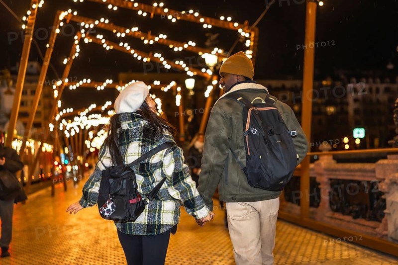 Young multiethnic couple walking on bridge with christmas lights, enjoying winter holidays traveling in europe