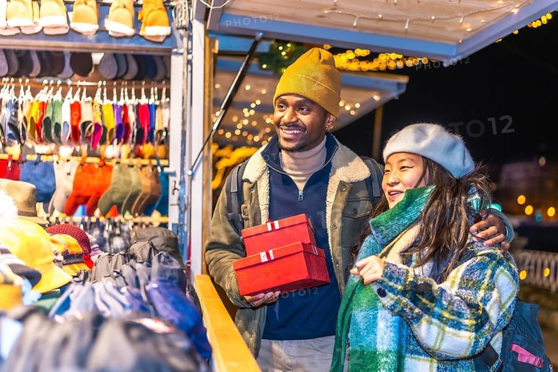 Smiling multiethnic couple holding christmas gifts while enjoying christmas market at night
