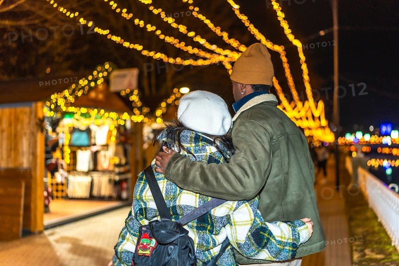 Young multiethnic couple walking hand in hand, sharing warm hugs amidst the glowing lights of a festive christmas market at night