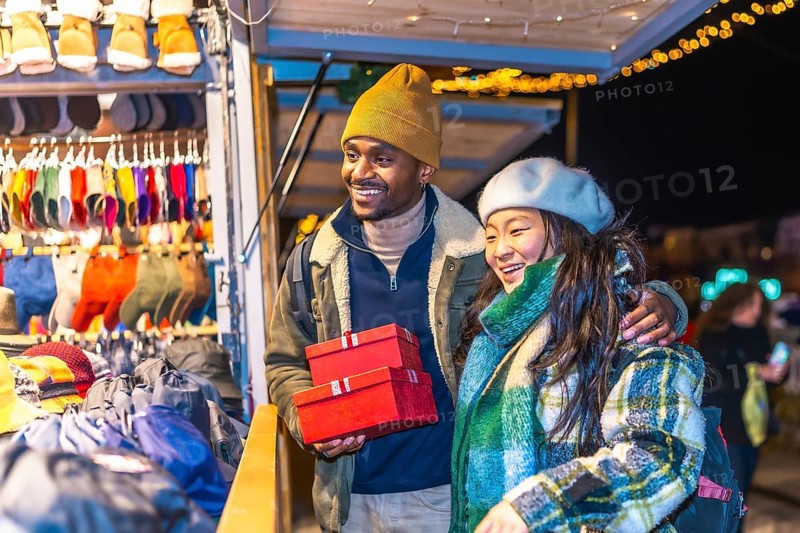 Multiethnic couple enjoying christmas shopping at a decorated christmas market, holding red gift boxes and smiling