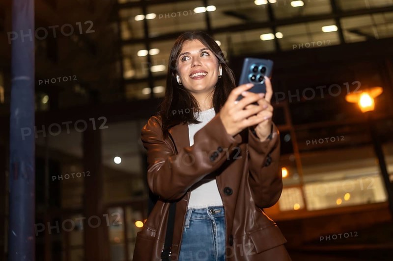 Young woman happily smiling and looking up while using her smartphone and wireless earbuds at night, capturing urban moments or communicating with friends in a city environment