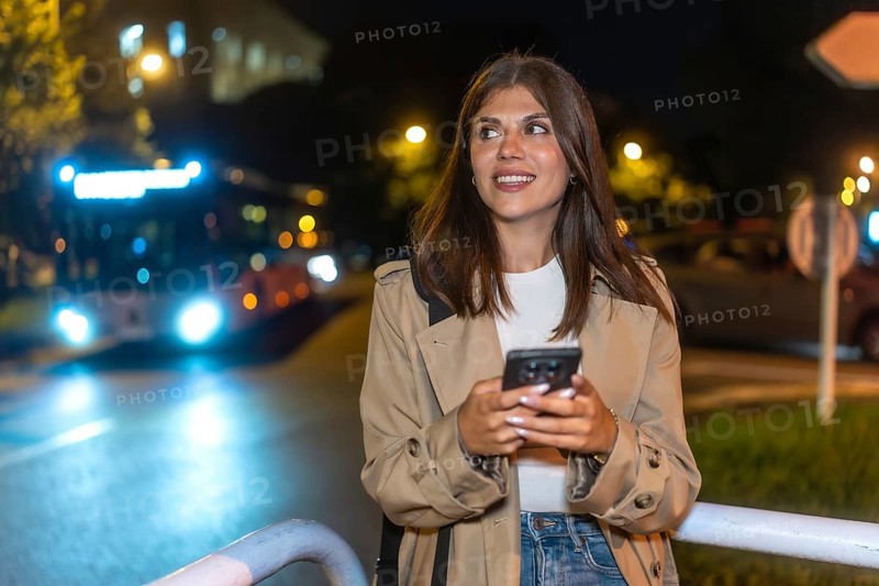 Young woman smiling while checking her mobile phone at a city bus stop at night, anticipating transportation or meeting friends in an urban environment