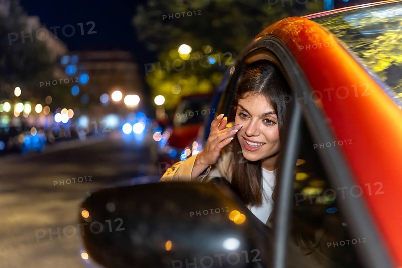 Young woman looking at herself in the car's side mirror, adjusting her makeup while parked on a city street at night, illuminated by urban lights and warm bokeh