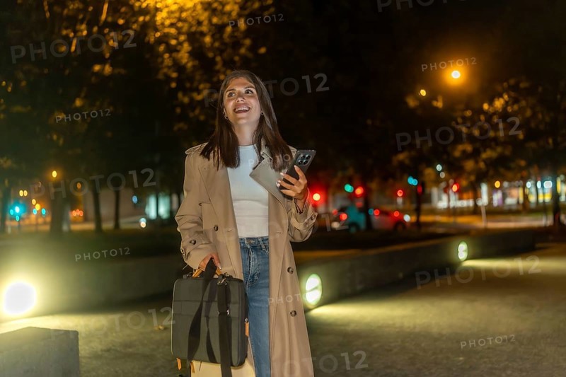 Woman in trench coat walking through an illuminated city park at night, smiling and confident while checking her smartphone, enjoying a peaceful urban evening stroll