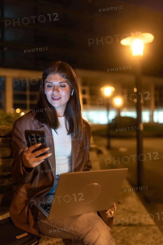 Young woman engaging in remote work and communication, looking at her smartphone while holding a laptop under the glow of urban street lights in a city environment