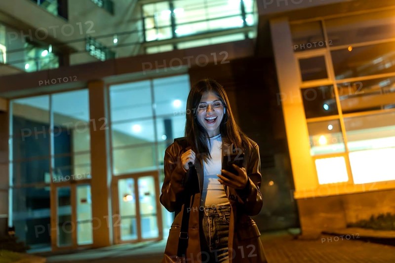 Young woman standing on an urban street at night, happily looking at her smartphone while holding a shoulder bag, with a modern building's illuminated facade in the background