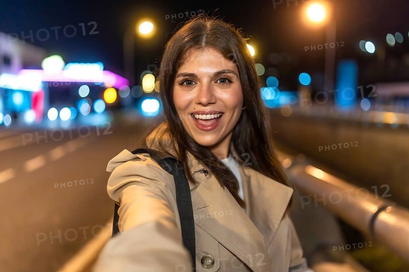 Young woman smiling and taking a selfie on a lively city night, bokeh street lights and light trails glowing behind her as she explores urban streets in a trench coat