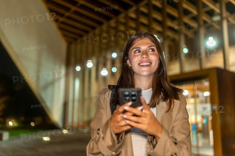 Young woman smiling while holding a smartphone and looking up, exploring a modern city street at night, experiencing technology and connectivity in an urban setting