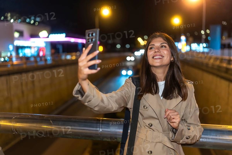 Young woman smiling and holding a smartphone, capturing a selfie against the bright, blurred lights of a busy urban street at night, enjoying personal connection