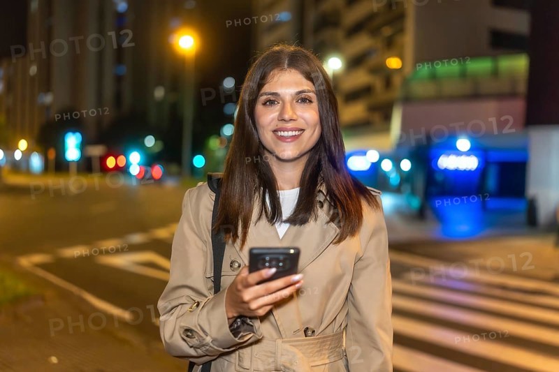 Woman smiling brightly and holding a smartphone, standing on a bustling city street at night with illuminated buildings and traffic lights creating a vibrant urban background