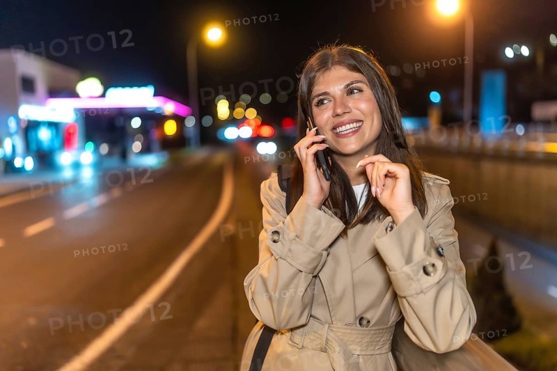 Young woman in a trench coat smiling as she talks on her mobile phone on a bustling city street at night, bathed in warm urban lights and soft bokeh background