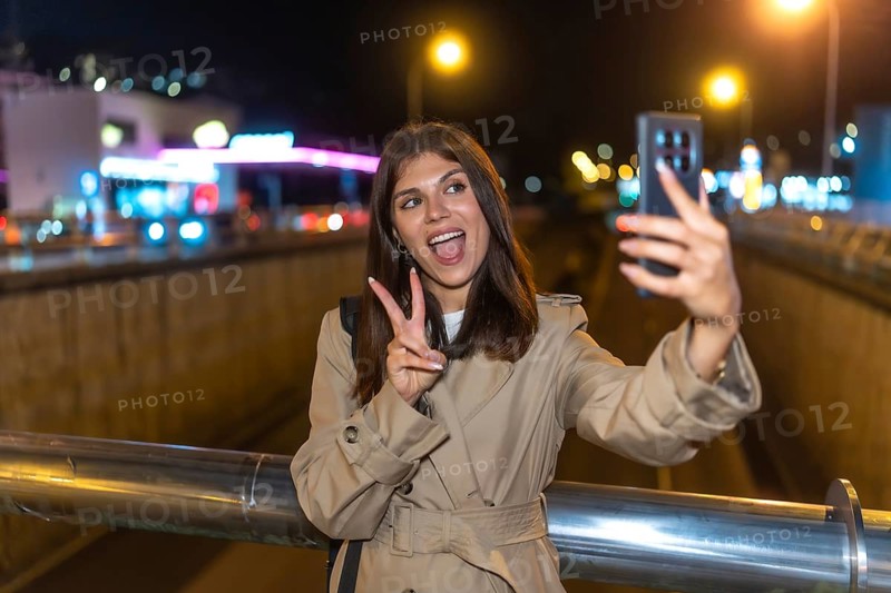 Young woman wearing a trench coat standing on a bridge, holding a smartphone and taking a self portrait, showing the peace sign with a happy expression during a vibrant city night