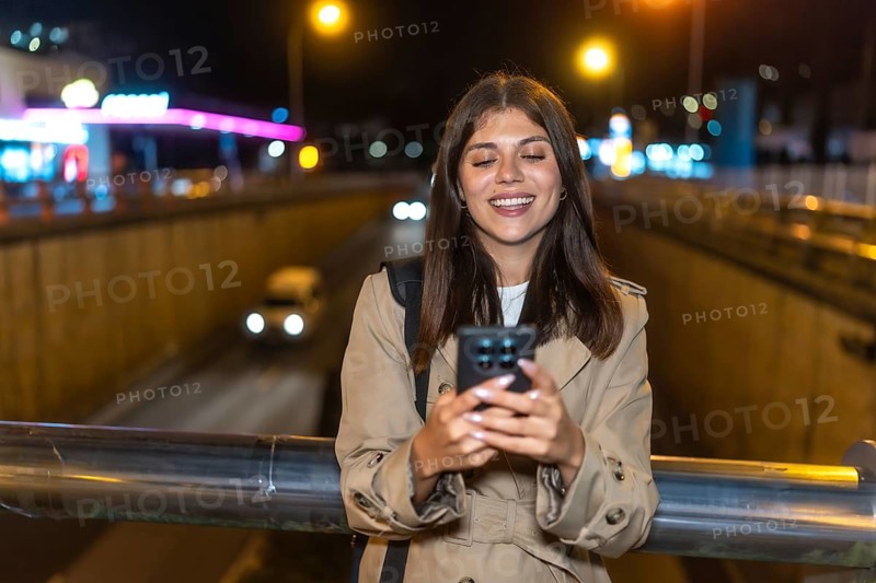 Young woman standing on a bridge at night, smiling as she uses her smartphone with city lights and blurred traffic creating bokeh urban background, casual and content