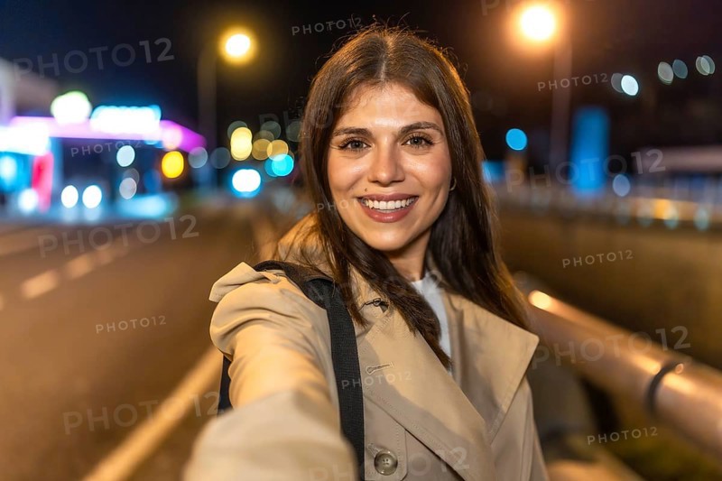 Young woman smiling and holding a smartphone, capturing a selfie portrait outdoors in an urban environment at night, surrounded by blurred city lights and a vibrant atmosphere