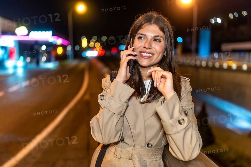 Young woman in a trench coat smiling happily while having a phone call, standing on a city street at night with blurred lights creating a vibrant urban background