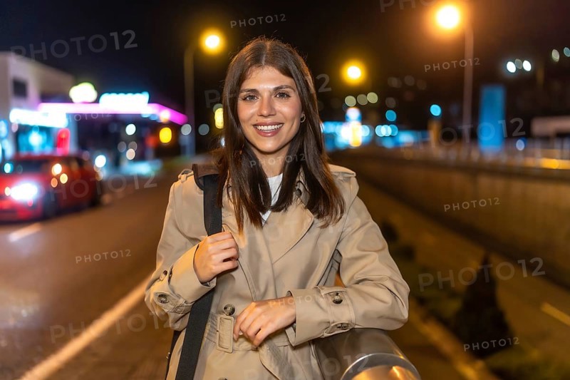 Young woman smiling while standing in a trench coat on an urban street at night, illuminated by city lights and a gas station in the blurred background