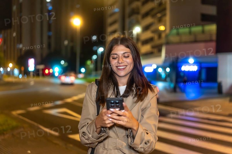 Young woman smiling with eyes closed, enjoying her smartphone on a city street at night, relaxed and content while texting, browsing social media amid glowing streetlights and bokeh