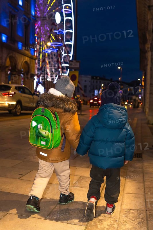 Two children holding hands and walking away from the camera on a sidewalk near a brightly lit ferris wheel at christmas time in bayonne, france