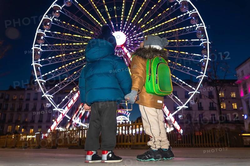 Two children holding hands are admiring a brightly lit ferris wheel at a christmas market in bayonne, france, creating a magical winter holiday scene