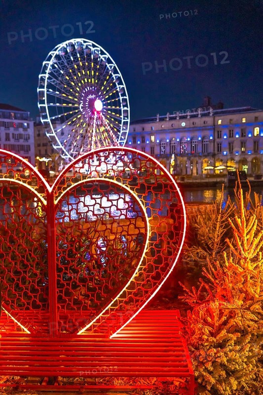 Illuminated heart shaped bench with small christmas trees and ferris wheel in the background, creating a romantic atmosphere in bayonne, france