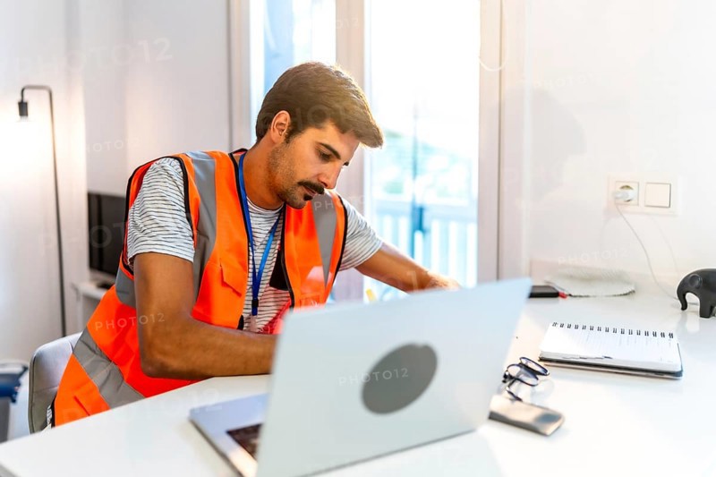 Focused engineer wearing a high visibility vest working from home, using a laptop and taking notes in a bright, modern home office