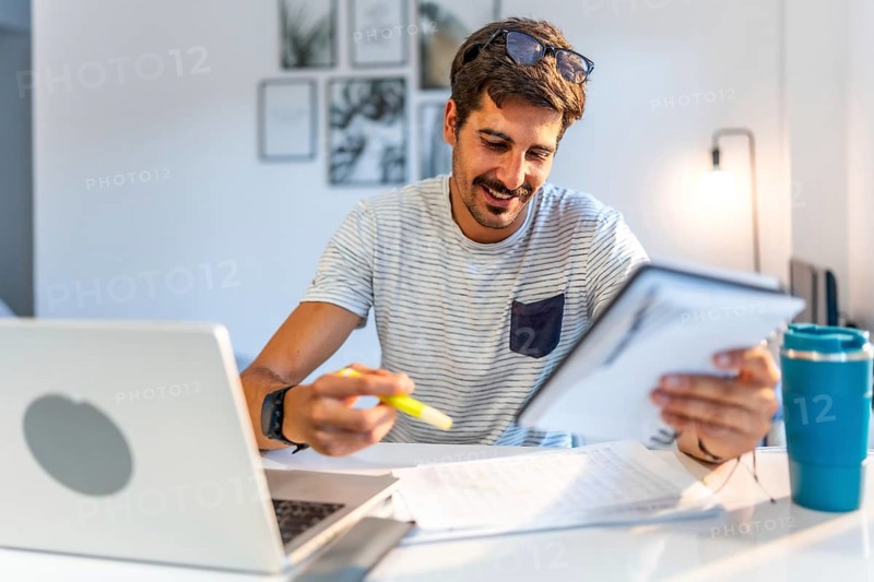Young male freelancer comparing information on his tablet with data on his laptop, working from his modern home office