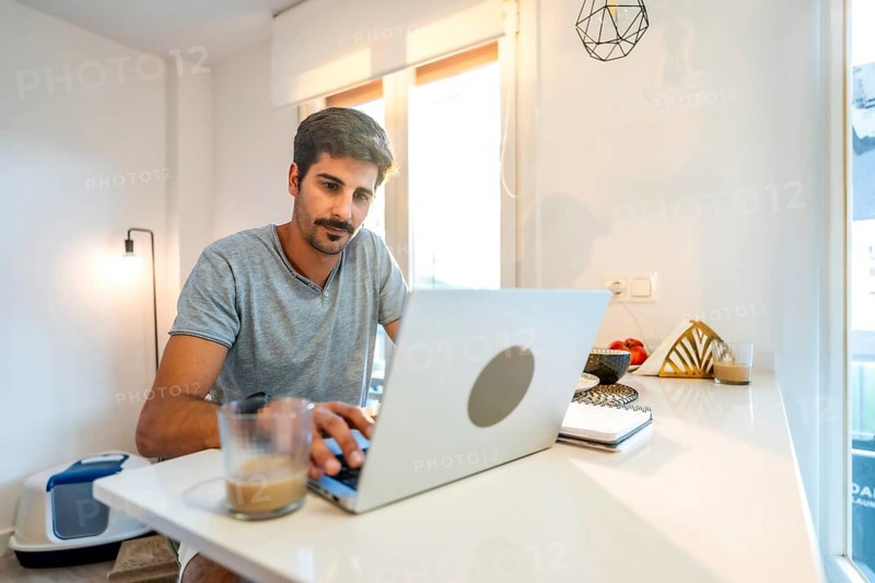 Young man concentrating on his laptop, enjoying a coffee while working remotely from his stylish kitchen