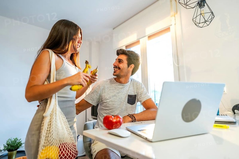 Happy young couple is eating bananas and planning next grocery shopping online using laptop in their modern kitchen