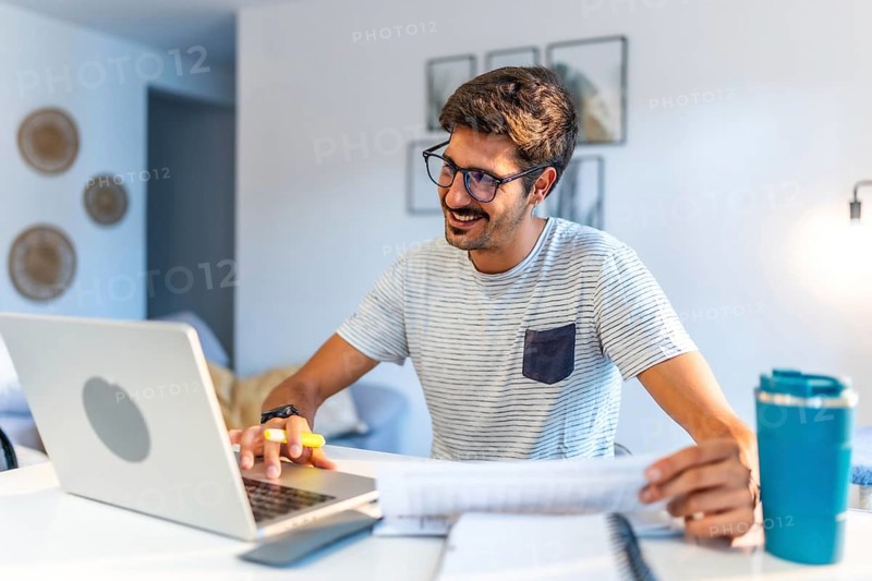 Young man working comfortably at home, engaging with a laptop and reviewing documents, smiling while enjoying a productive day