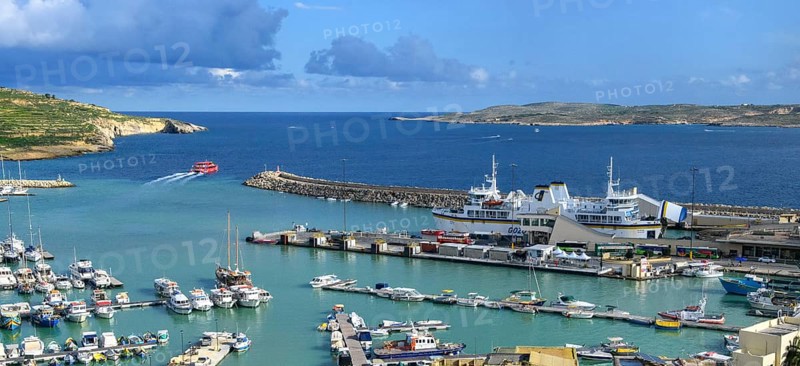 View of harbour basin of Gozo Mgarr Harbour with small boats yachts yachts, in the middle of the picture jetty for ferries from to Valletta island Malta and moored ferry car ferry, in the background part of Comino island, Mgarr, Gozo, Malta
