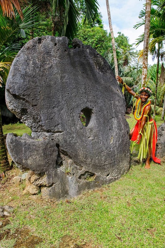 Local boy in traditional traditional costume from Yap Island as size comparison next to oversized large huge coins made of stone real means of payment in stone money bank stone money bank in jungle of Yap Island, Yap Island, Yap State, Caroline Islands, FSM, Federated States of Micronesia, Australia, Oceania