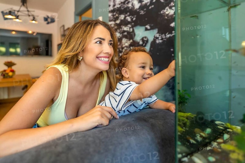 Happy mother and her baby son lying on the sofa, enjoying a delightful moment together while watching the colorful fish in the aquarium