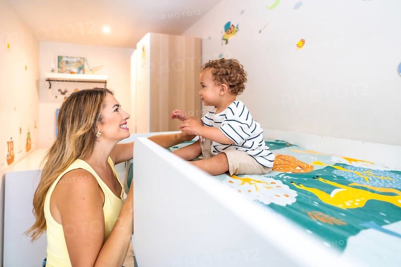 Smiling mother looking at her cute baby sitting on a bunk bed in a decorated bedroom, happy family concept