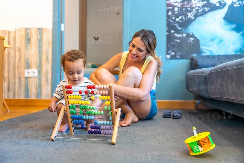 Happy mother watching her baby boy playing with a colorful abacus, developing early math skills in a playful home environment