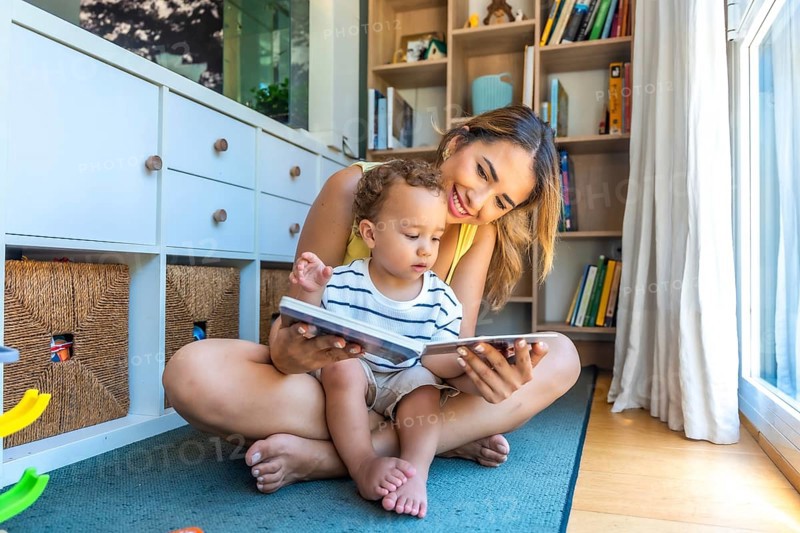 Smiling mother and baby son sharing joyful moments while reading books together, fostering a love for literacy in their cozy playroom