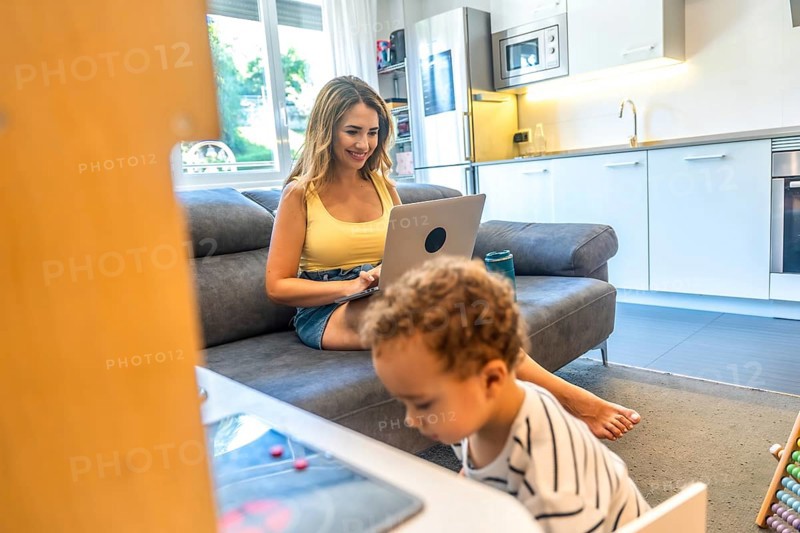 Woman telecommuting on her laptop while her baby plays nearby in a modern living room, blending work and family life seamlessly