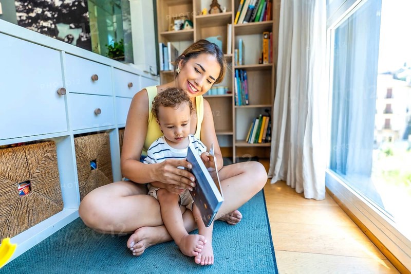 Happy mother and her little son reading a book together, sitting on the floor in the living room