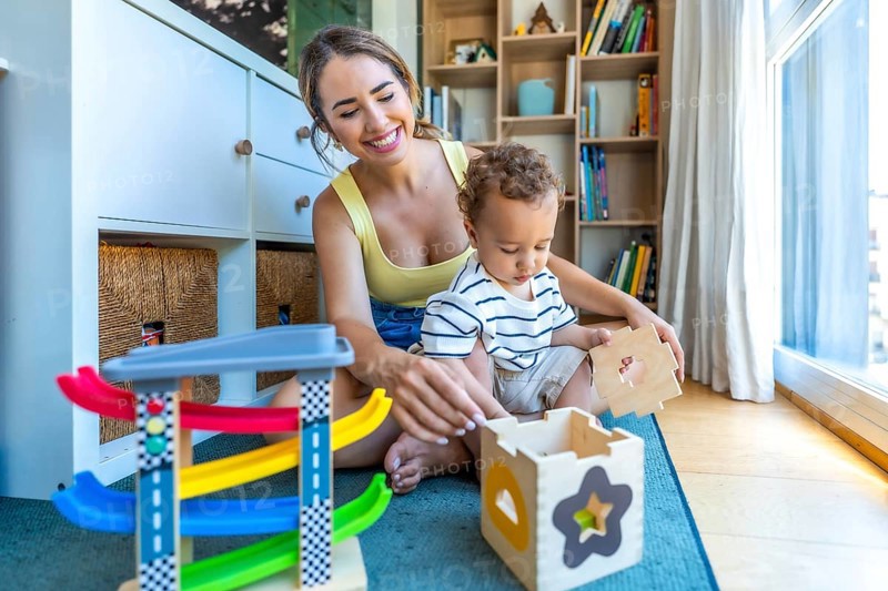 Happy mother and son are sitting on the floor playing with a wooden shape sorter and colorful toy car ramp in a cozy kids room