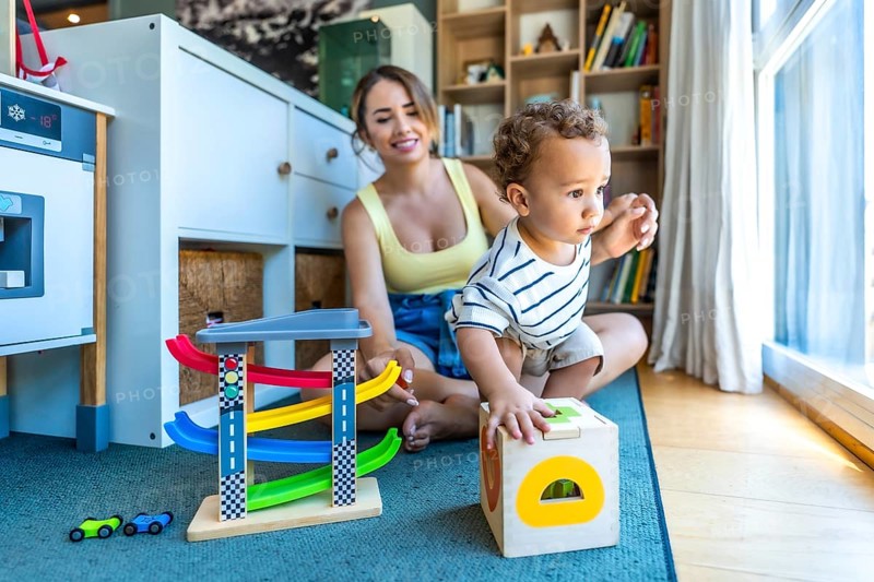 Happy mother watching her toddler son playing with educational wooden toys in a modern living room
