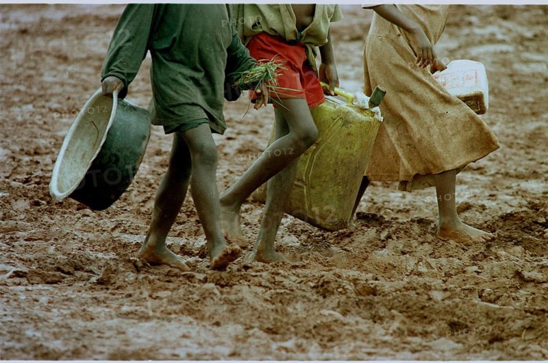 RWA, Benaco camp, 19.05.1994 -- Refugee children wander through the muddy paths of the Benaco camp to get food and water. -- Rwandan refuggee children walk a muddy road for their daily food and water supply at the Benaco camp.