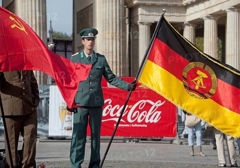 DEU Germany Berlin Actor wearing NVA uniform poses with the GDR flag in front of a Coca-Cola commercial. Scene on the eve of the 20th anniversary of German reunification on the festival mile in Berlin. Germany Berlin Actor in GDR Army Uniform Poses with GDR Flag in Front of a Coke Banner. Scene from the public party one day ahead of the celebration marking the 20th anniversary of the German reunification